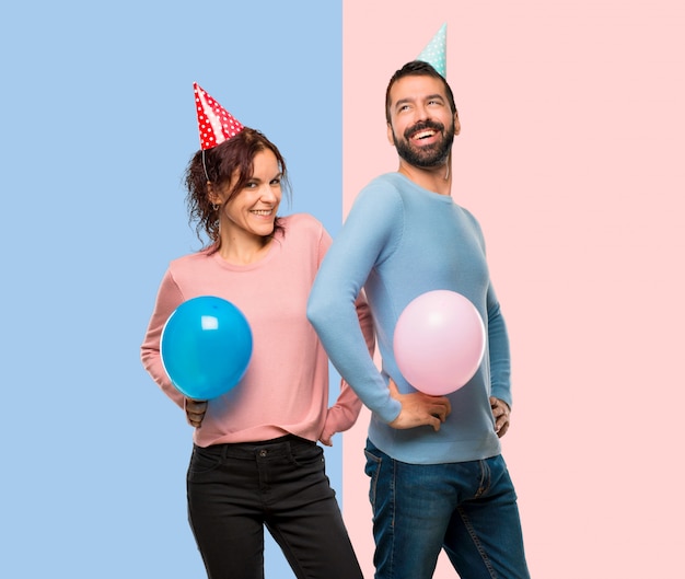 Coppia con palloncini e cappelli di compleanno in posa con le braccia in anca e ridendo