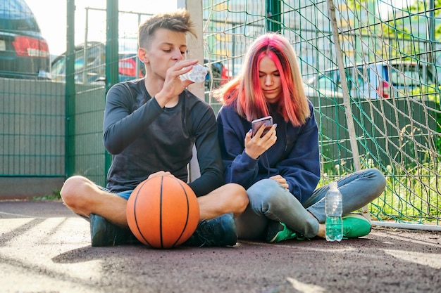 Coppia di adolescenti sul campo sportivo di strada con palla, stile di vita, ragazzo e ragazza di 16 anni che parlano, bevendo acqua dalla bottiglia guardando lo schermo dello smartphone