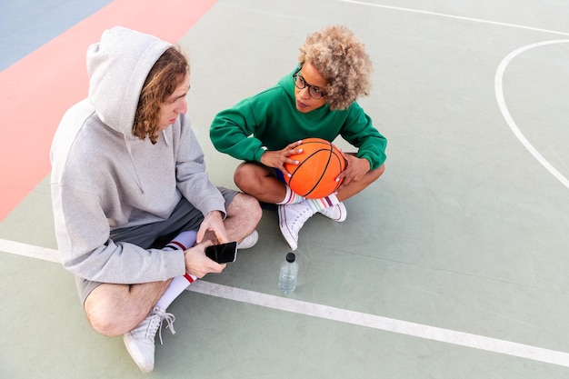 Coppia di amici che chiacchierano e si divertono seduti in campo dopo una partita di basket in un parco cittadino concetto di amicizia e sport urbano in strada copia spazio per il testo