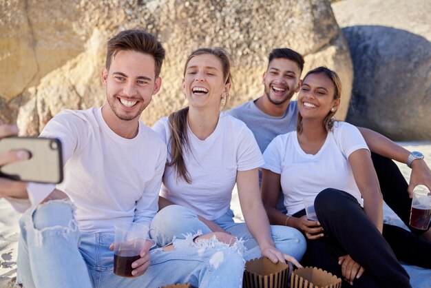 Coppia di amici selfie e drink durante un picnic sulla spiaggia con un sorriso per un aggiornamento sui social media mentre sono in vacanza in estate Uomini e donne con il telefono al doppio appuntamento alle rocce per divertimento e libertà all'aperto