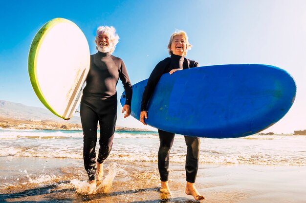 Coppia di anziani in spiaggia con mute nere in possesso di un surftable pronto per andare a fare surf in spiaggia - persone attive mature e pensionate che fanno attività felici insieme nelle loro vacanze o nel tempo libero