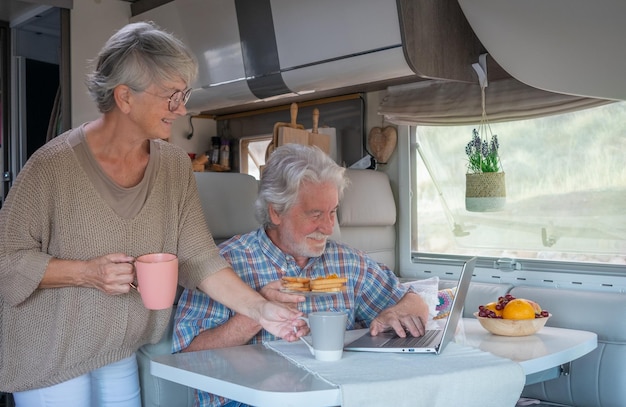 Coppia di anziani in vacanza di viaggio all'interno di un camper godendo la colazione insieme Coppia senior caucasica felice nella vita di un furgone mentre naviga sul computer portatile Luce brillante dalla finestra