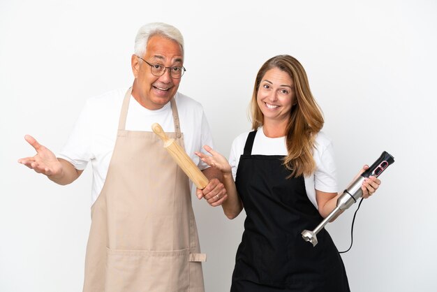 Coppia di chef di mezza età isolati su sfondo bianco che hanno dubbi mentre si alzano le mani e le spalle