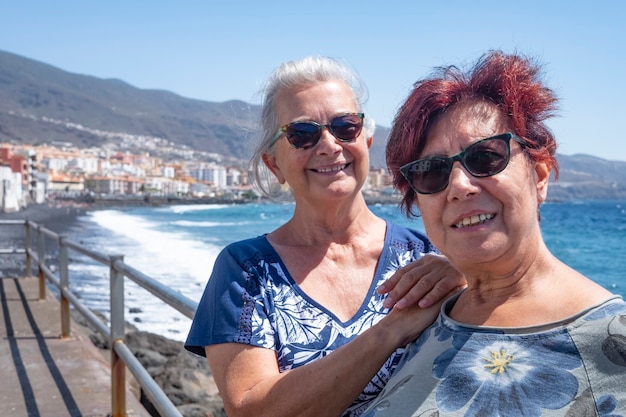 Coppia di due amiche che si godono un'escursione in mare appoggiata alla ringhiera del lungomare in una giornata di sole - vento, onde e vista sul mare a Tenerife