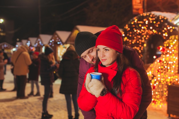 Coppia di giovani innamorati alla fiera di Natale. Uomo in piedi dietro la donna che tiene la sua vita, donna che tiene una tazza di bevanda calda nelle sue mani, distogliendo lo sguardo. Luci di Natale della fiera invernale su uno sfondo