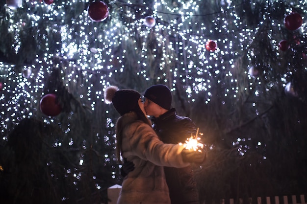 Coppia di innamorati con una scintillante luce bengala che si bacia vicino a un albero di Natale