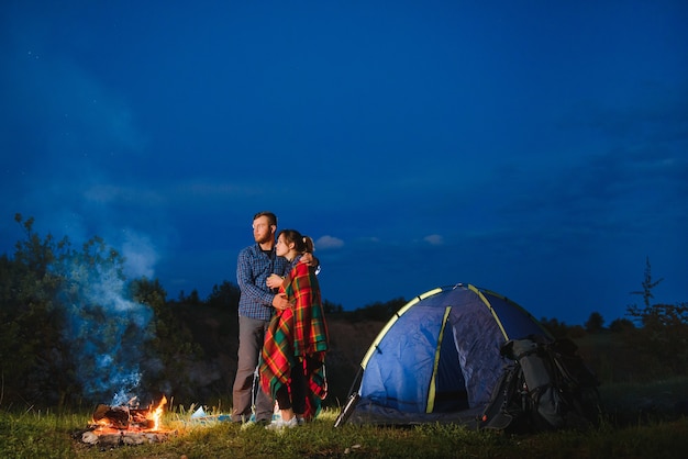 Coppia di innamorati escursionisti che si godono a vicenda, in piedi accanto al fuoco di notte sotto il cielo serale vicino a alberi e tenda