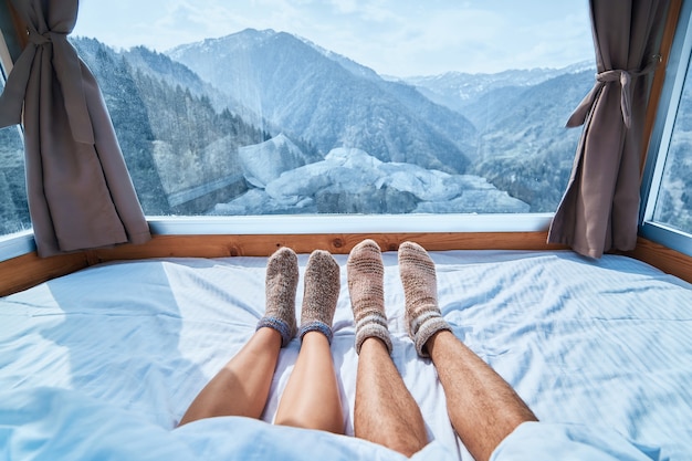 Coppia di innamorati in calzini caldi lavorati a maglia sdraiati su una camera da letto morbida e accogliente con vista sulle montagne innevate in inverno