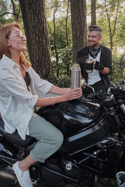 Coppia di mezza età che beve il tè seduto su una motocicletta che viaggiano insieme su una strada forestale