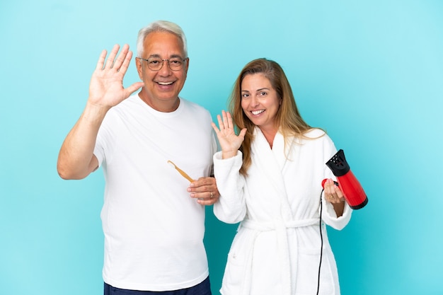 Coppia di mezza età che tiene asciugatrice e spazzolino da denti isolati su sfondo blu salutando con la mano con espressione felice