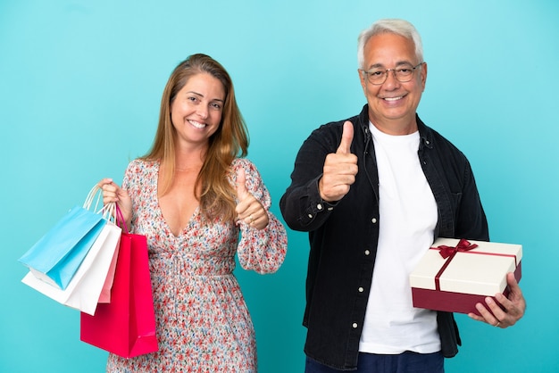 Coppia di mezza età con borsa della spesa e regalo isolato su sfondo blu che fa un gesto di pollice in alto perché è successo qualcosa di buono