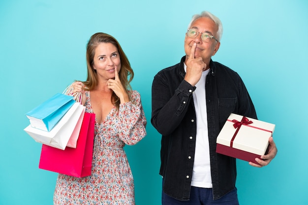 Coppia di mezza età con borsa della spesa e regalo isolato su sfondo blu sorridente con un'espressione dolce