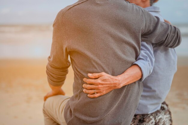 Coppia di pensionati in una sera d'estate guarda il mare, donna che abbraccia il marito in spiaggia al tramonto.