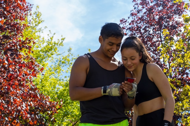 Coppia di sportivi che consultano il cellulare durante il loro allenamento di boxe all'aperto nel parco in una giornata di sole mostrando la loro felicità con volti sorridenti