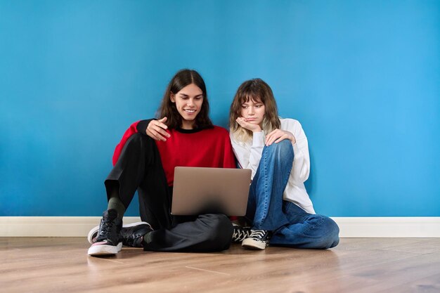 Coppia di studenti adolescenti che usano il computer portatile seduto sul pavimento su sfondo blu