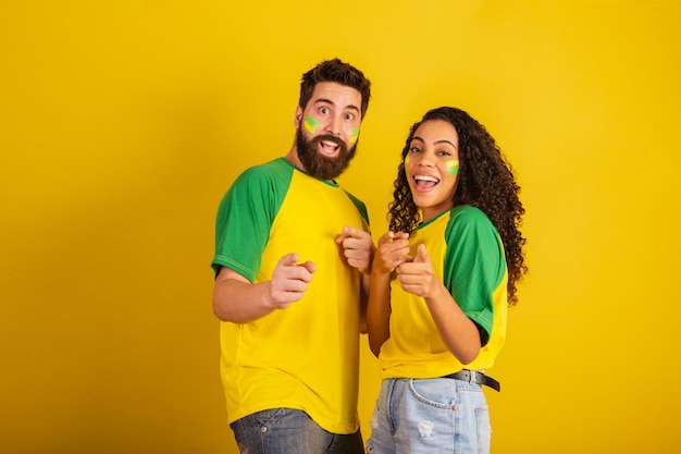 Coppia di tifosi di calcio brasiliani vestiti con i colori della nazione donna nera uomo caucasico che punta alla telecamera