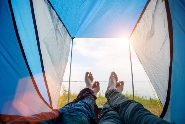 Coppia di turisti irriconoscibili che giace all'interno della tenda blu, osservando il bellissimo paesaggio fluviale. Vacanze di famiglia. Vista dall'interno della tenda, attraversata a piedi nudi di innamorati. Concetto di viaggio ed escursionismo.