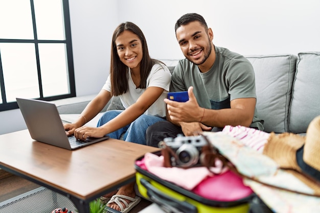 Coppia di uomini e donne latini che sorridono fiduciosi pagando per viaggiare utilizzando il computer portatile e la carta di credito a casa