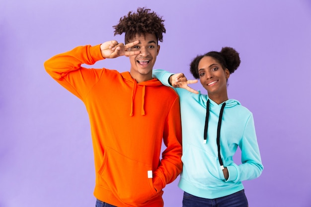 coppia divertente in abiti colorati, sorridente e mostrando il segno di pace, isolato sopra il muro viola