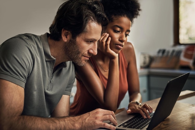 Coppia eterogenea alla ricerca di un nuovo appartamento in affitto a casa sul proprio laptop seduti insieme nella loro cucina a casa loro Uomo e donna in una relazione di matrimonio interrazziale che fanno shopping online