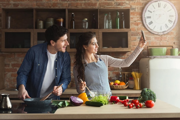 Coppia felice che cucina la cena e si fa selfie nella loro cucina soppalcata a casa. Preparare insalata di verdure e fotografare per i social network, copiare lo spazio