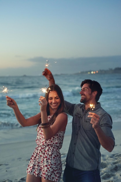 Coppia felice che tiene le stelle filanti per celebrare il capodanno sulla spiaggia al tramonto con fuochi d'artificio scintillanti