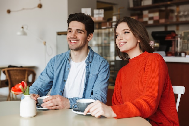 Coppia felice donna e uomo che sorride e che osserva da parte, mentre riposa nella caffetteria e beve caffè o tè insieme