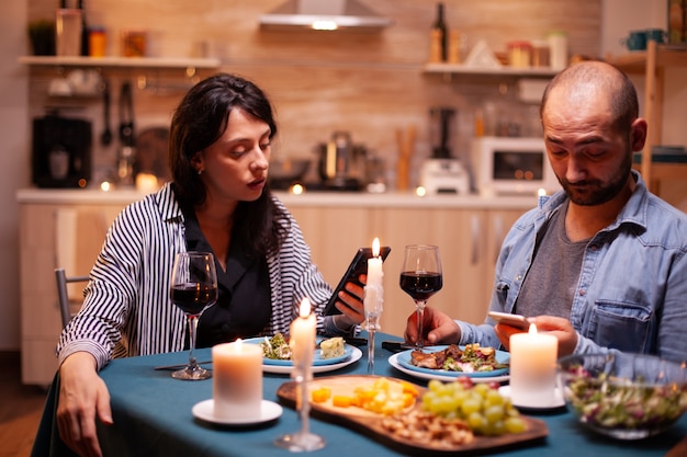 Coppia felice guardando video su smartphone in cucina durante l'anniversario. Adulti seduti al tavolo in cucina navigano, cercano, usano smartphone, internet, festeggiano l'anniversario.