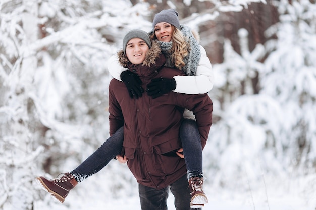 Coppia felice innamorata in inverno in un bosco innevato, una ragazza cavalca sul retro del suo amato ragazzo giocando e sorridendo