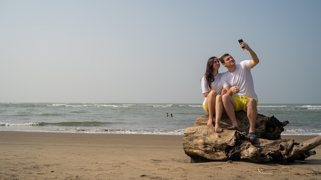 Coppia felice seduta su legni vicino al mare Coppia di innamorati che si abbracciano e si fanno selfie mentre si siede sul log durante l'appuntamento sulla spiaggia contro il mare ondeggiante e il cielo senza nuvole