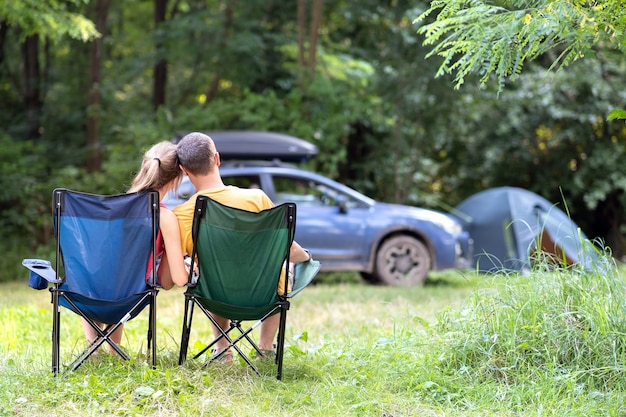 Coppia felice seduto su sedie in campeggio abbracciando insieme a un'auto e una tenda sullo sfondo.