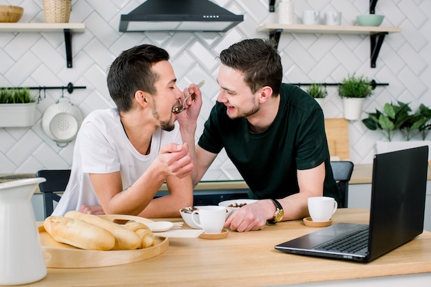 Coppia gay maschio seduto al tavolo e fare colazione nella loro cucina. Giovane bello in maglietta nera che alimenta il suo partner. Coppia gay