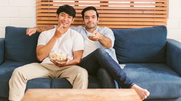 Coppia gay seduta sul divano con popcorn e godendosi la visione di contenuti in tv nel soggiorno LGBT caucasica e asiatica che vivono insieme Diversità dell'amicizia gay