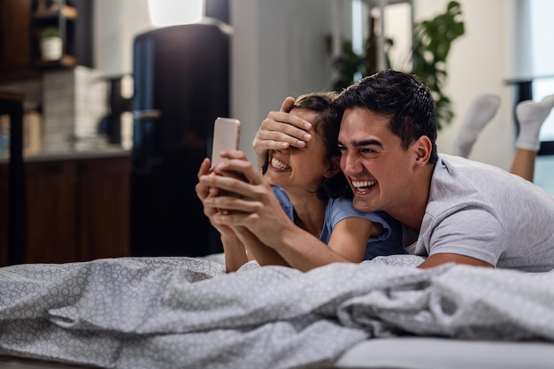 Coppia giocosa che si fa selfie con il cellulare e si diverte in camera da letto L'uomo sta coprendo gli occhi della donna