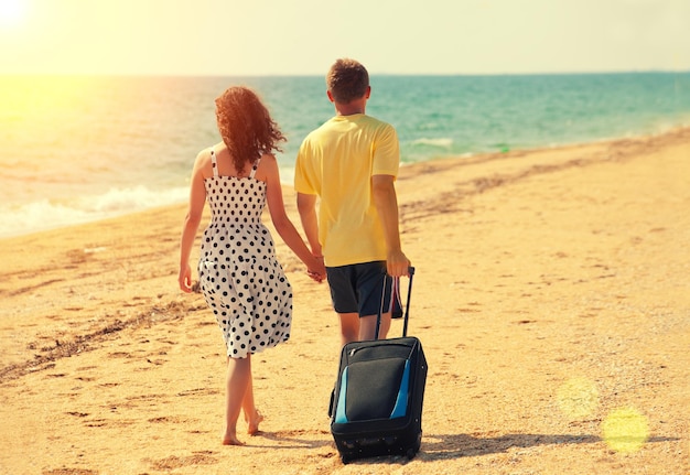 Coppia giovane con borsa da viaggio che si tiene per mano camminando sulla spiaggia torna alla fotocamera