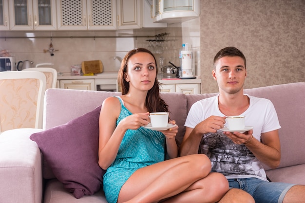 Coppia giovane con tazze di caffè o tè seduti insieme sul divano e guardare la televisione con espressioni vuote