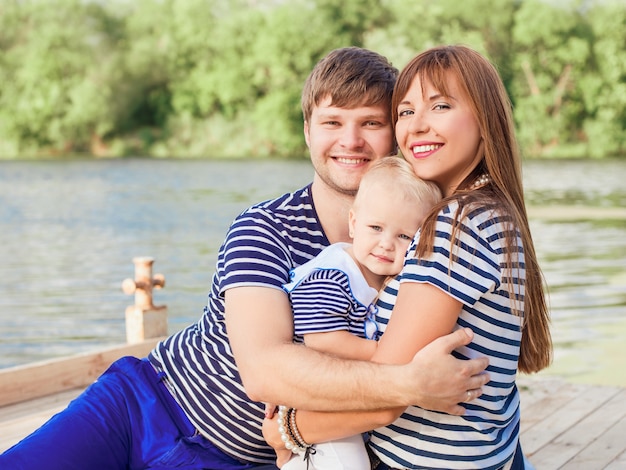 Coppia giovane donna e uomo in magliette a righe seduti e tenendo in braccio il loro bambino in riva al fiume
