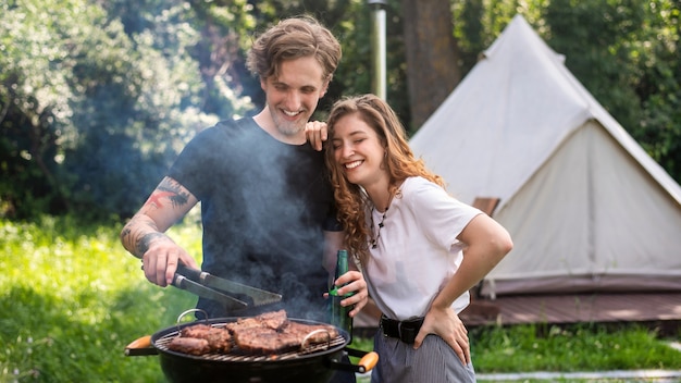 Coppia giovane friggere carne alla griglia e bere birra, ridendo. Verde intorno. Glamping