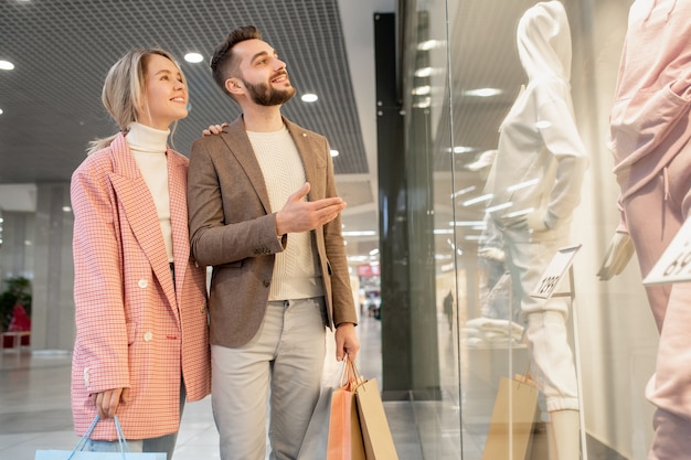 Coppia giovane guardando il manichino in tuta dietro la vetrina del negozio durante i loro acquisti nel centro commerciale