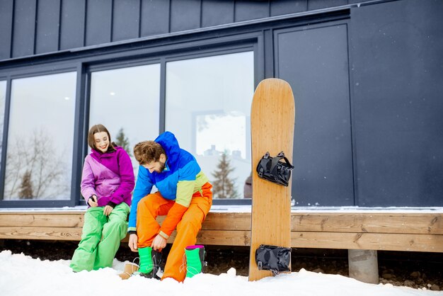 Coppia giovane in tute da sci che si preparano per lo snowboard seduti sulla terrazza della casa moderna in montagna