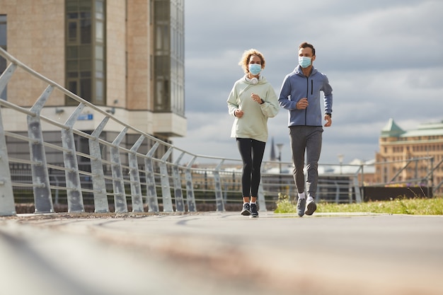 Coppia giovane indossando maschere protettive fare jogging insieme la mattina lungo la strada