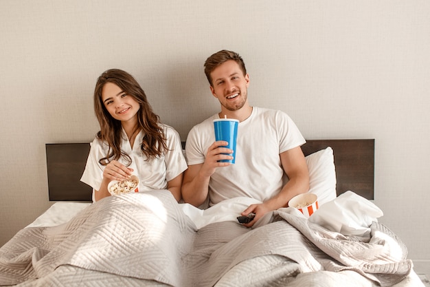 Coppia giovane nel letto. Il bei uomo e donna sorridenti stanno mangiando il popcorn e stanno guardando la TV insieme in camera da letto