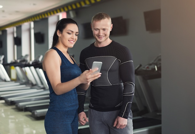 Coppia giovane utilizza lo smartphone in palestra. Donna atleta che guarda le foto sullo smartphone con il suo allenatore, facendo una pausa dopo l'allenamento, copia spazio