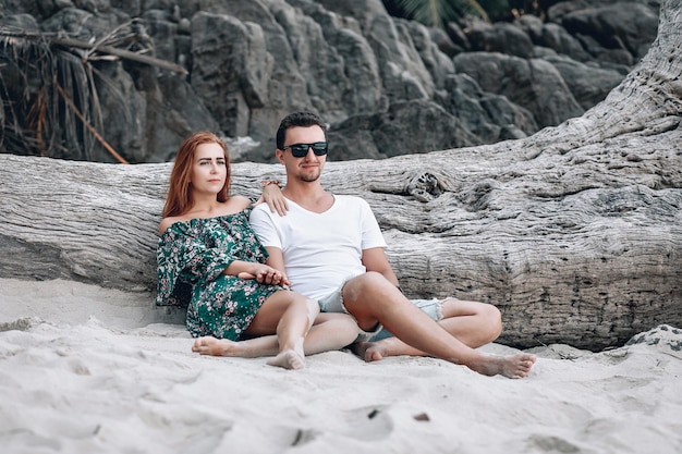 Coppia hipster sulla spiaggia, ragazza con i capelli rossi in abito floreale verde e uomo con stoppie, seduto vicino al vecchio grande albero nudo senza foglie. Phuket. Tailandia