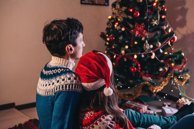 Coppia in amore seduto accanto a un albero di Natale, con indosso il cappello di Babbo Natale e abbracci.