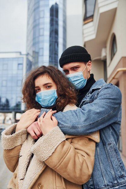 Coppia in maschere protettive che si abbracciano in città vicino all'edificio degli affari in tempo di quarantena Concezione del coronavirus