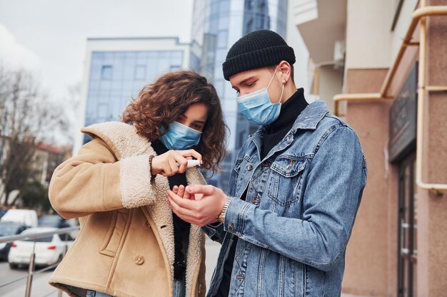 Coppia in maschere protettive che usano antisettico all'aperto in città vicino all'edificio degli affari in tempo di quarantena Concezione del coronavirus