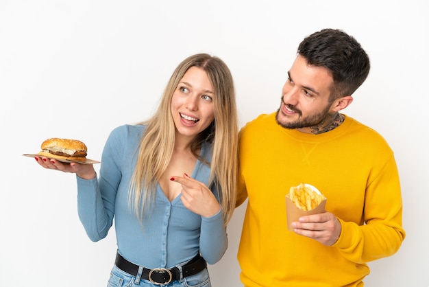 Coppia in possesso di hamburger e patatine fritte su sfondo bianco isolato che presenta un'idea mentre guarda sorridendo verso