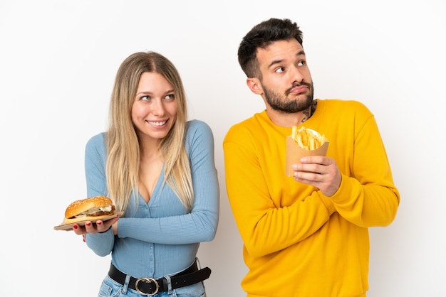 Coppia in possesso di hamburger e patatine fritte su sfondo bianco isolato facendo gesti di dubbi mentre si sollevano le spalle