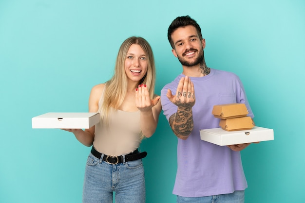 Coppia in possesso di pizze e hamburger su sfondo blu isolato che invita a venire con la mano. Felice che tu sia venuto